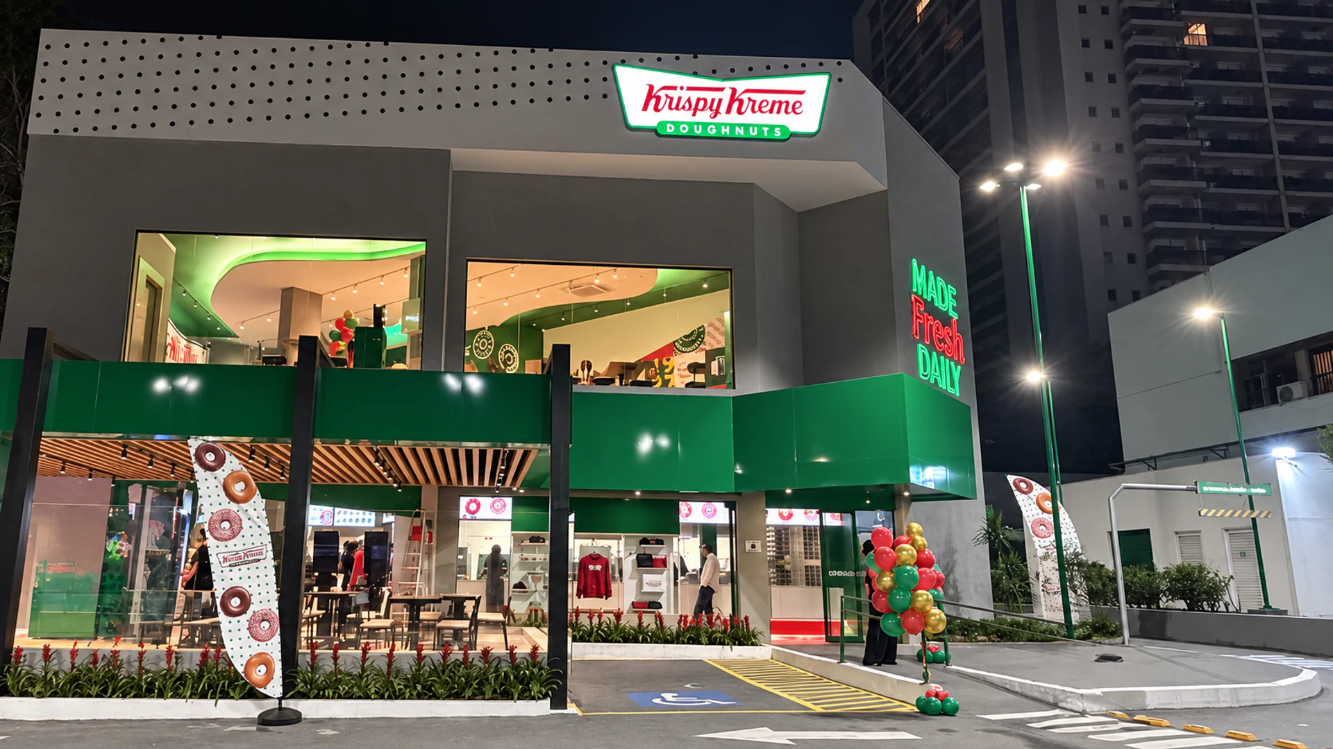 Fotografia real da fachada da Krispy Kreme na avenida dos Bandeirantes, com dois pavimentos, grandes vãos de vidro e marquise verde iluminada