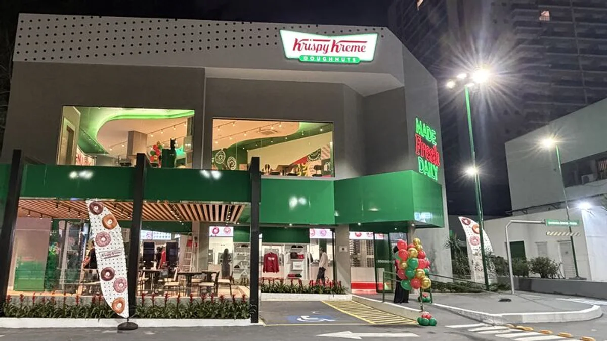 Fotografia real da fachada da Krispy Kreme na avenida dos Bandeirantes, com dois pavimentos, grandes vãos de vidro e marquise verde iluminada